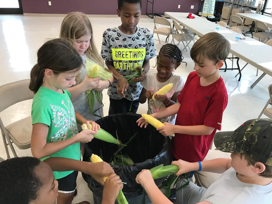 Children stand around a large garbage can while they shuck the husks and silks from ears of corn. 