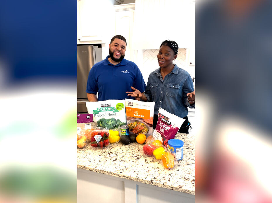 A man and woman stand behind a kitchen counter filled with fruits and vegetables.
