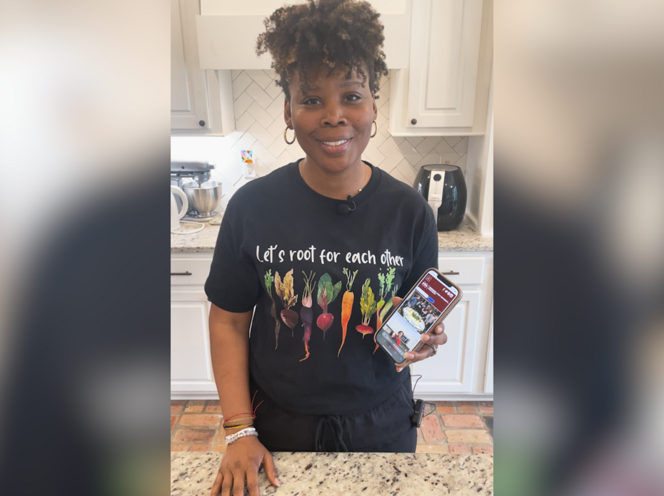 A woman stands in kitchen with a cell phone displaying a web page.