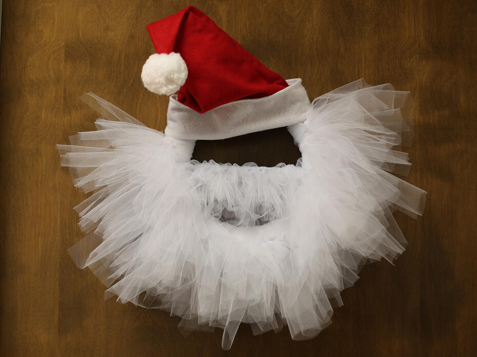 Wreath with Santa hat and white tulle shaped to look like a beard hanging on a wooden door.