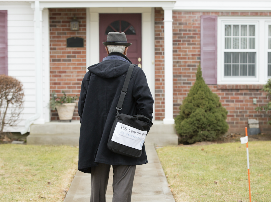 A census worker wearing a black hat and coat walking up to a house.