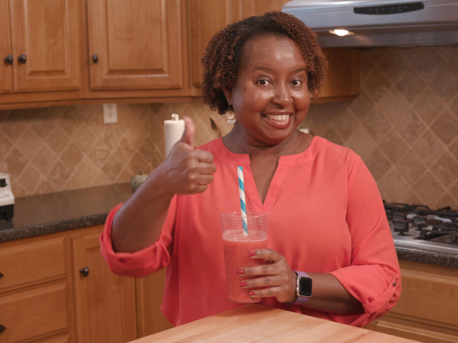 A lady in a kitchen holds a smoothie and gives a thumbs up.
