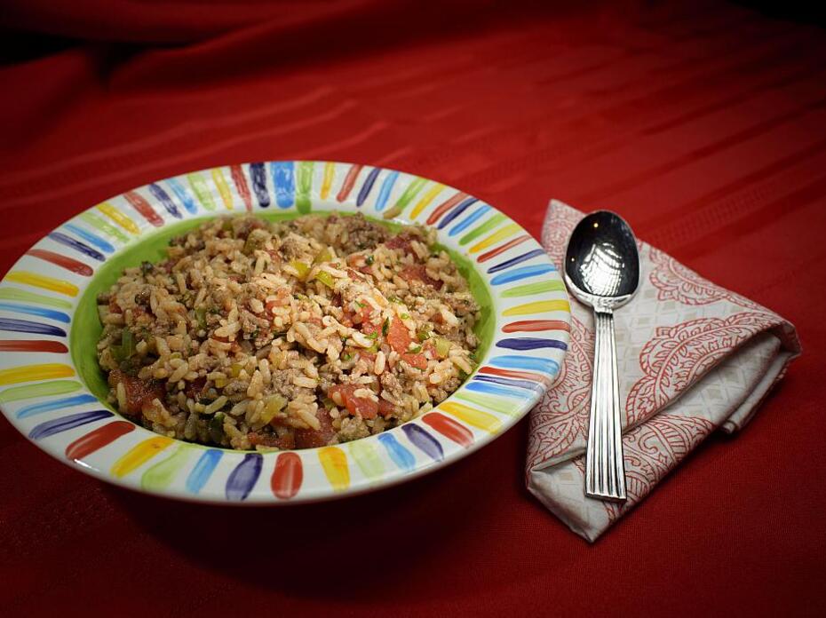 A bowl of Italian ground beef and rice in a rainbow-striped bowl sits on a red tablecloth with a red and white cloth napkin and a spoon.