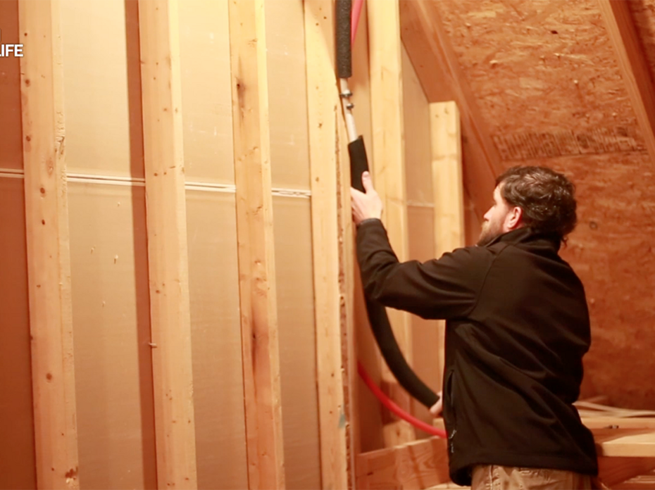 A man fits insulation around water pipes in an unfinished attic of a home.