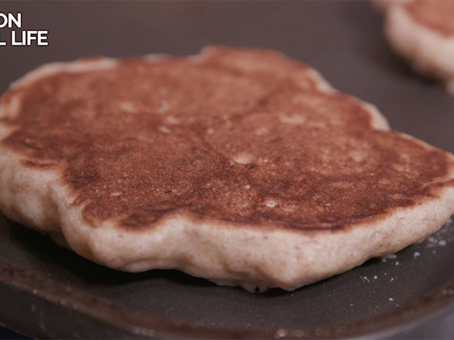 A close-up of a golden brown whole-grain pancake on a nonstick griddle, with parts of two other pancakes blurred in the background. 
