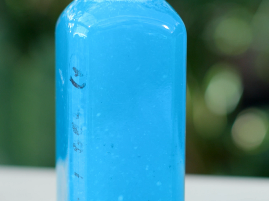 Close-up of a portion of a clear bottle filled with bright blue liquid that is water-soluble fertilizer dissolved in water, against a blurred dark green background.