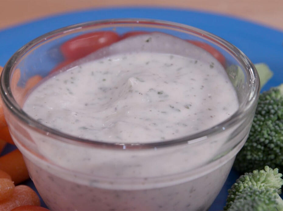 Homemade ranch dressing in a clear glass bowl atop a turquoise blue plate, surrounded by baby carrots, broccoli florets, and grape tomatoes.