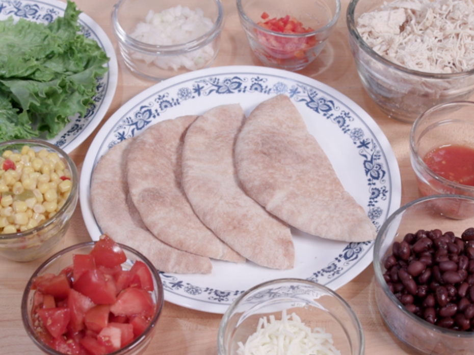 An array of ingredients in glass bowls, including romaine lettuce, onion, diced red pepper, Mexicorn, diced tomato, black beans, salsa, cooked diced chicken breast, and mozzarella cheese, surround a blue and white plate bearing four halves of whole wheat pita bread.