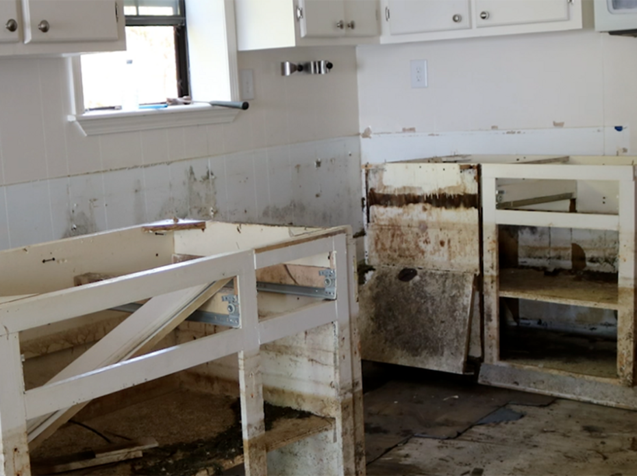 A kitchen with white walls and cabinets shows severe flood damage, including mold damage, on floors and cabinets with doors and drawers removed.