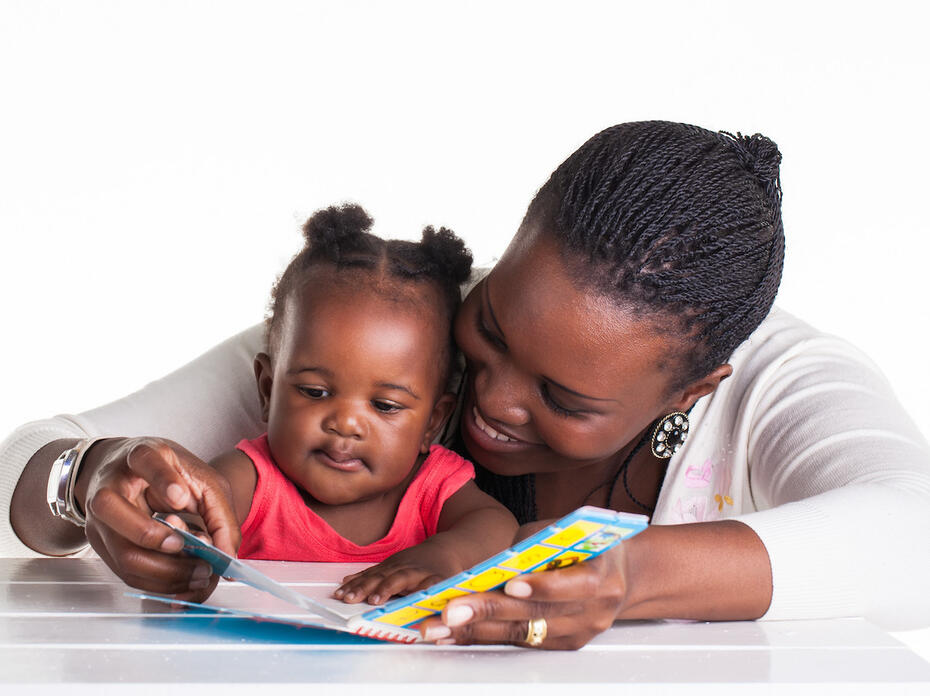 An African American woman with braided hair and wearing a white sweater shares a board book with her infant daughter who wears a coral shirt. 