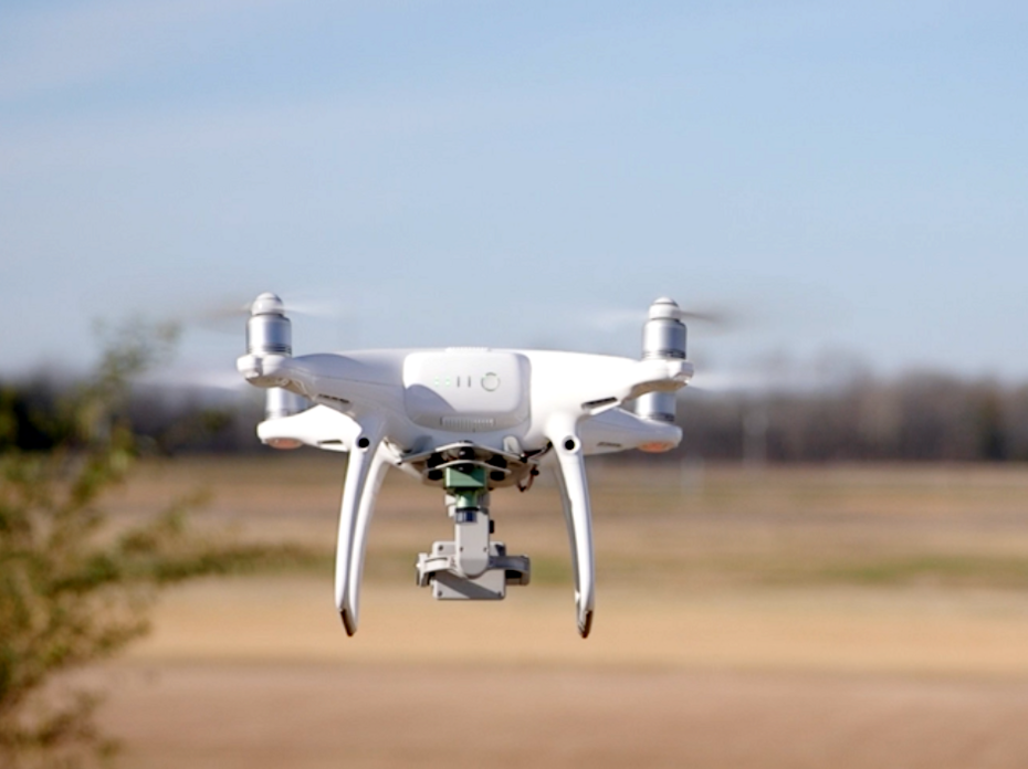 A close up shows a drone hovering above an open field. 