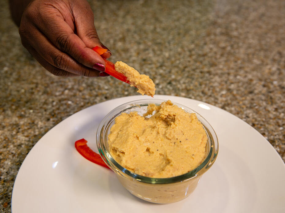 A hand is shown dipping a bell pepper strip into a bowl of garlic chickpea dip.