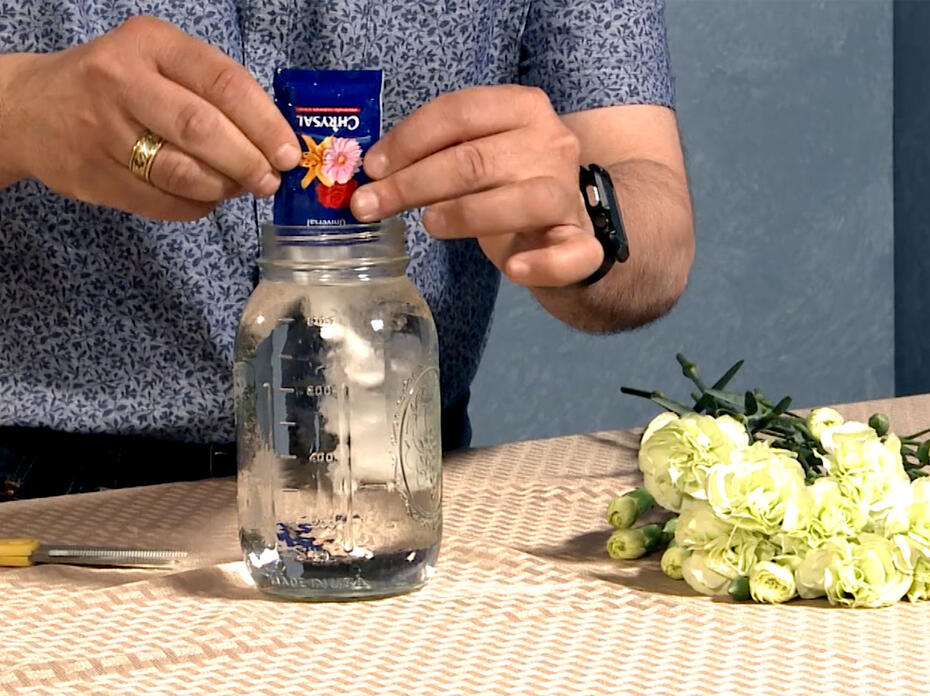 A man in a blue shirt adds a packet of flower food to a clear glass quart jar. White flowers lay beside the jar on a cloth-covered table.