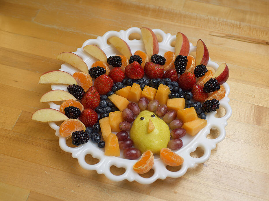Several rows of fruit including apple slices, tangerine segments, blackberries, blueberries, cantaloupe, strawberries, grapes and a pear are arranged on a white platter in the shape of a turkey.