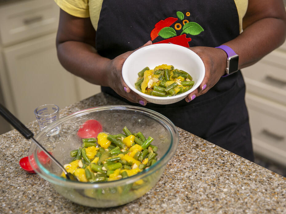 A bowl of peachy green beans sits on the kitchen counter with a woman holding a serving size in a separate bowl.
