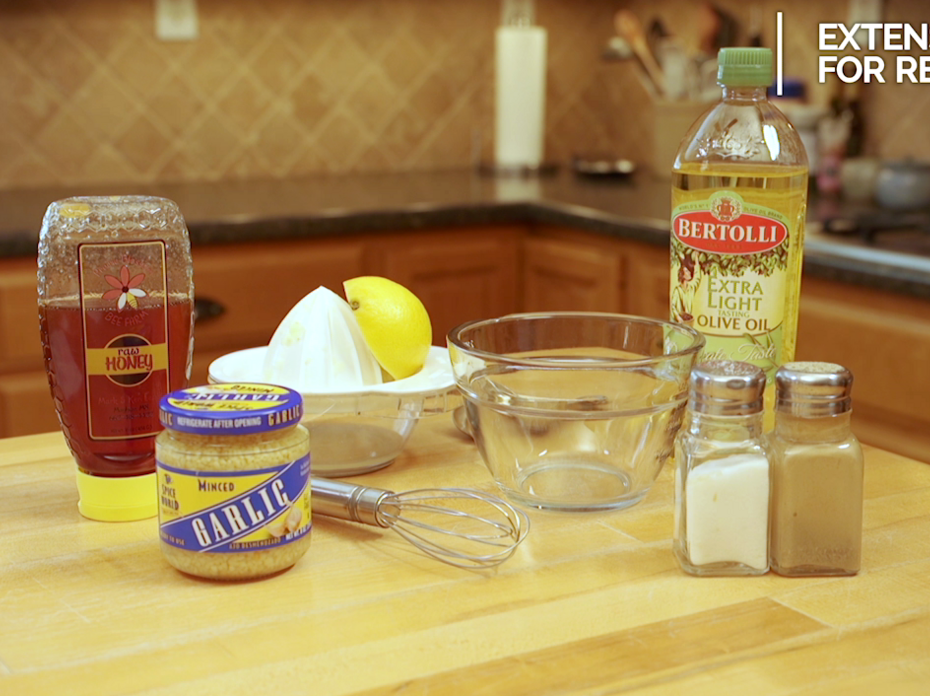 The ingredients and equipment to make honey lemon vinaigrette are displayed on a kitchen table, including honey, olive oil, half of a lemon, salt, pepper, minced garlic, a small glass bowl, a stainless steel whisk, and a citrus juicer. 