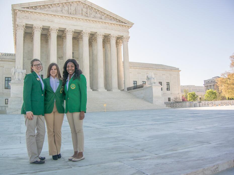 Three female teenagers wearing green blazers and khaki pants standing in front of the Supreme Court Building in Washington, D.C.