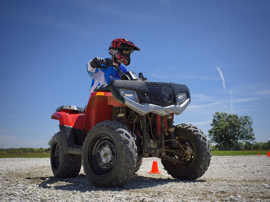 A teenage boy in a blue and white long-sleeved shirt and protective gear, including a red and black helmet and black gloves, rides a red ATV around orange cones during a safety class. (Kevin Hudson)
