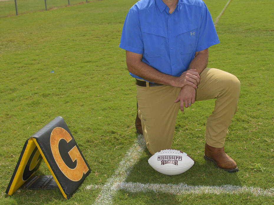 A man kneels on a football practice field beside a football.