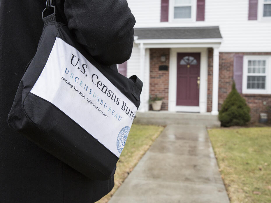 A person carrying a black bag marked with census information approaches the front door of a house.