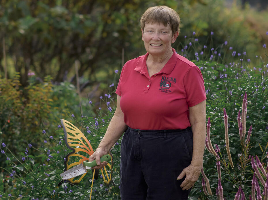 A woman holding a gardening tool stands in a green landscape.