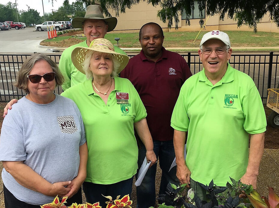 A group of five adults stand in front of some plants.