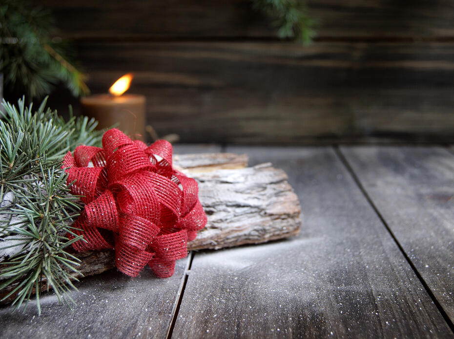 A red bow rests on a log with evergreen foliage in front of a candle.