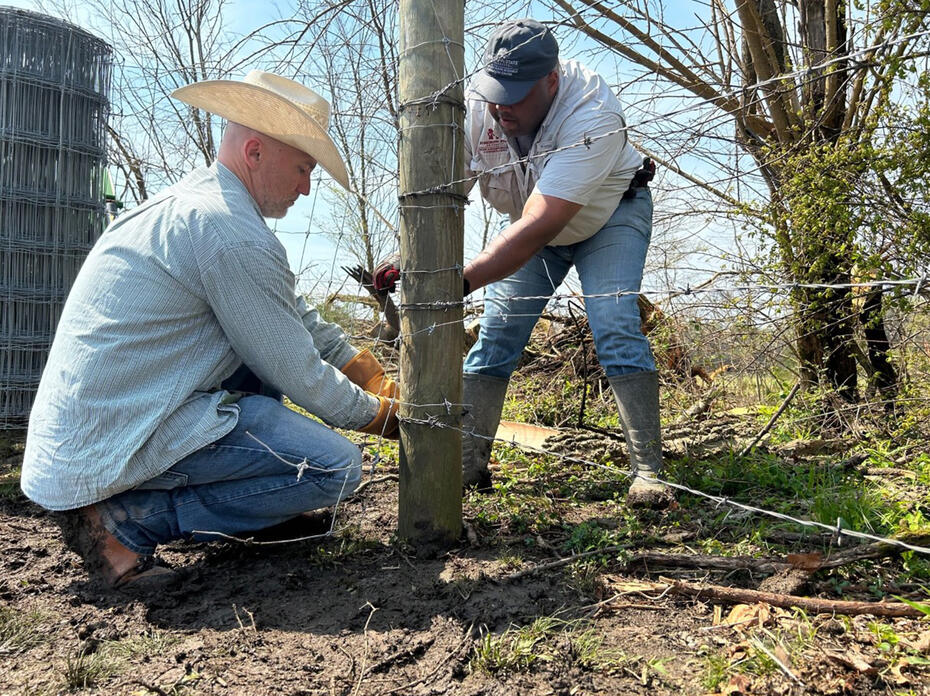 Two men tie barbed wire around a fence post.