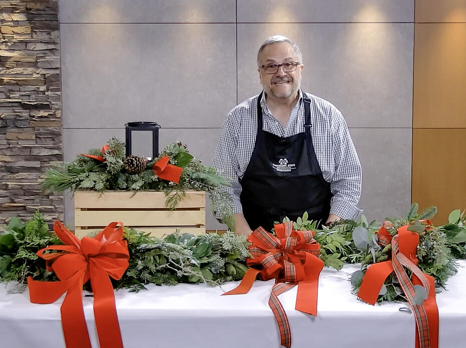 A man stands in a studio with a completed velvet bow, garland, door swag, and table centerpiece.