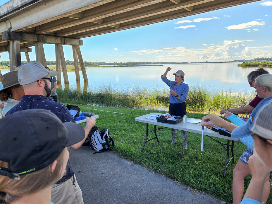 A woman stands at a table under a bridge while others look on.