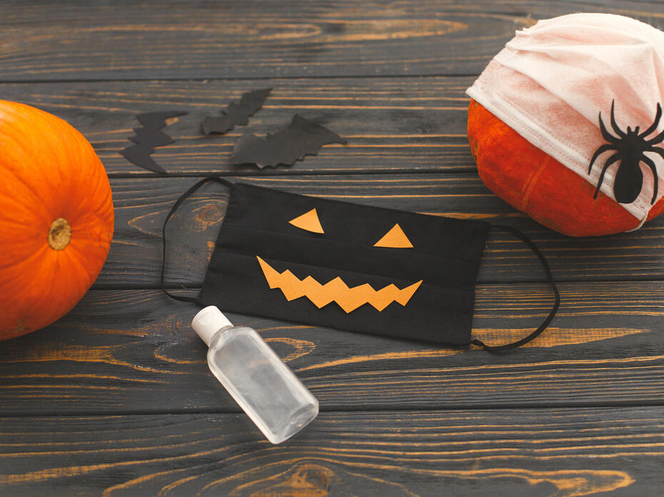 A black jack-o-lantern mask, bottle of sanitizer, and two pumpkins on a wooden backdrop.