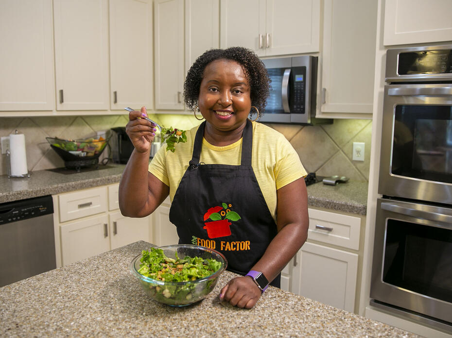 A woman in a kitchen smiles and holds a forkful of salad up for the camera.