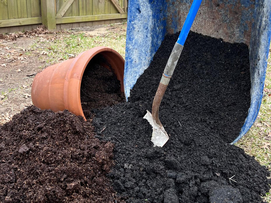 A shovel stands among soil from a wheelbarrow and a pot.