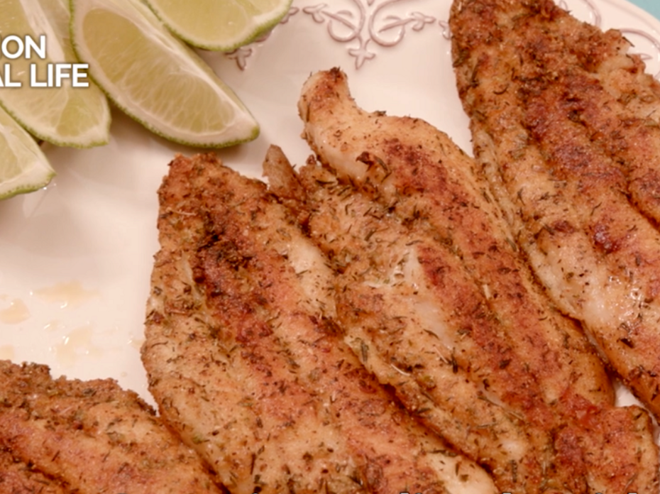 Four browned, seasoned catfish fillets on a white plate garnished with lime wedges.