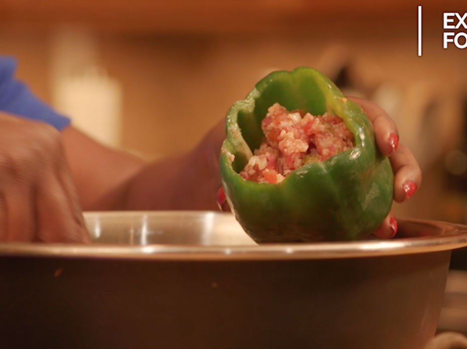 Hands spoon a ground meat and riced cauliflower mixture into a raw green bell pepper held over a stainless steel bowl. 