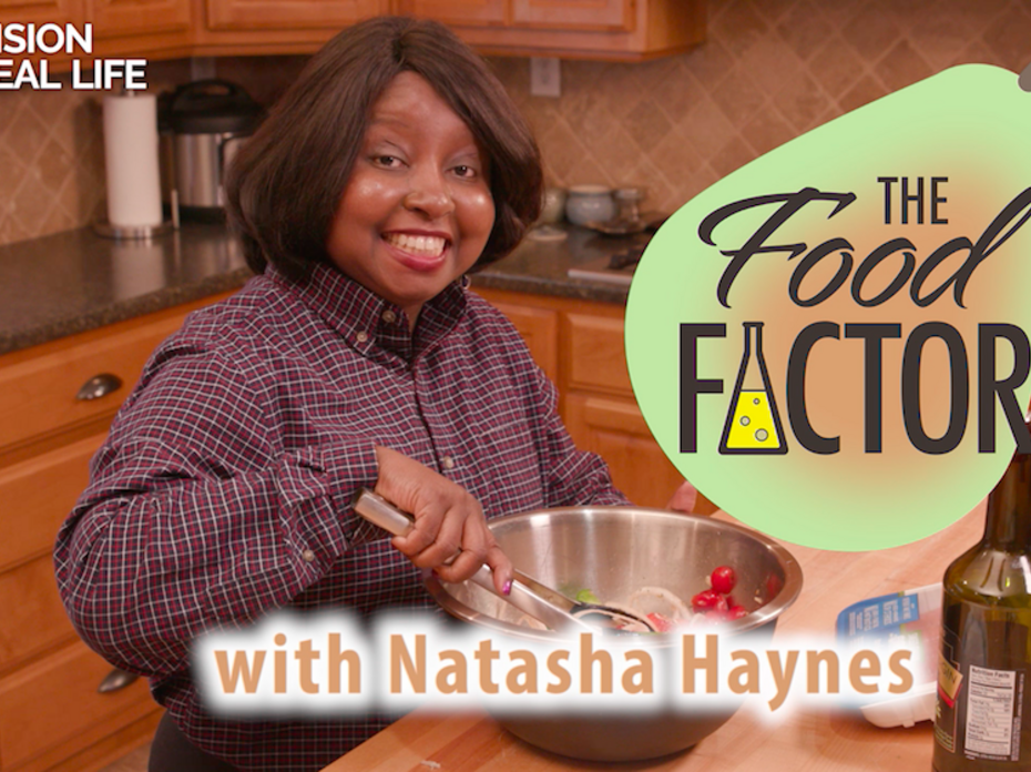 Natasha Haynes, an African American female wearing a plaid dress shirt stands in a kitchen and uses tongs to mix food ingredients in a stainless steel bowl. A tall bottle of olive oil is on the table. 