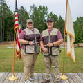 Two people, smiling and wearing 4-H Shooting Sports vests flanked by flags.