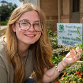 A person smiling, holding a plant in front of a sign listing, “Pearl River County 4-H Jr. Master Gardener Project.”