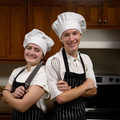 Two young people wearing chef's hats, with one holding a knife and one holding a whisk, standing back to back and smiling.