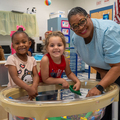 A smiling adult with two smiling children at a bead and building station.