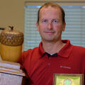 A man wearing a collared red shirt stands holding a large wooden trophy with a large wooden acorn on top in one hand and a plaque in the other.