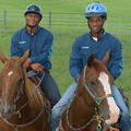 Two boys sit on two horses facing the camera. Both boys are wearing blue shirts and safety helmets.