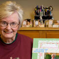 An elderly woman holds a scrapbook page while sitting behind a table full of several more pages.