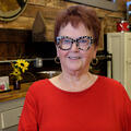 An elderly woman wearing animal print glasses and a bright red shirt stands smiling. Photo credit: Kevin Hudson