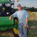 A man wearing a blue t-shirt and blue jeans leans against a green tractor.