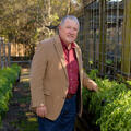 A gray-haired man with white goatee wears a brown blazer over a red polo with blue jeans and brown shoes. He stands in a dirt path flanked on both sides by parsley, about waist-high.