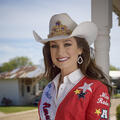A young woman wearing a white cowboy hat and a red embroidered blazer stands on a wooden deck.