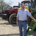 A man wearing a blue collared shirt rests his hand on a green tractor with several red tractors parked behind him.