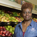 A woman wearing a blue collared jacket stands in the fruit aisle of a grocery store.