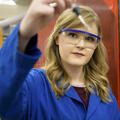 A blonde young woman wearing a blue lab coat and safety glasses holds a glass dropper up to the camera.
